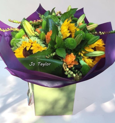Sunshine Meadow - A vibrant and uplifting arrangement bursting with warm golden tones and fresh greenery, wrapped in rich purple paper for a striking contrast. This hand-tied bouquet has a natural, garden-inspired feel, making it a cheerful gift perfect for brightening someone’s day or celebrating a special moment.