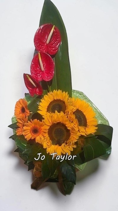 Tropical Sunset Statement Arrangement - A bold and eye-catching design featuring rich, warm tones contrasted with glossy greenery and a striking vertical accent. This contemporary arrangement has a sculptural, tropical feel, making it perfect for adding drama and vibrant energy to any space or special occasion.